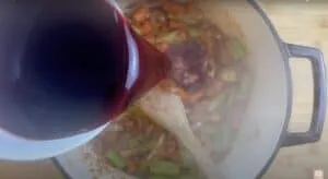 Pouring red wine into a pot with vegetables for cooking a savory dish.